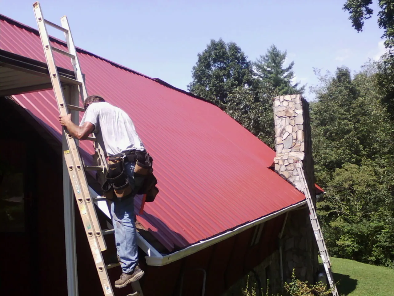 Roofer on a ladder beside a red metal roof and stone chimney — Soffit & Fascia Repair by Roofing Brothers in Dammeron Valley